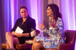 Two individuals seated and engaged in conversation under purple and blue lighting, with a small table and flower bouquet between them, one holding a paper and the other gesturing.