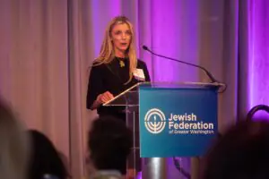 Speaker at a podium with a microphone, presenting in front of purple and gray curtains, with a sign reading "Jewish Federation of Greater Washington."