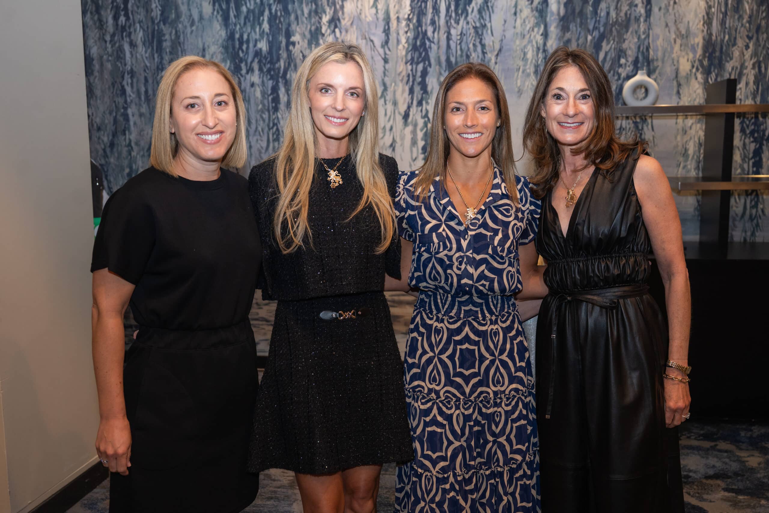 Four women standing side by side in front of a blue and white abstract background, dressed in various styles of black and patterned dresses.