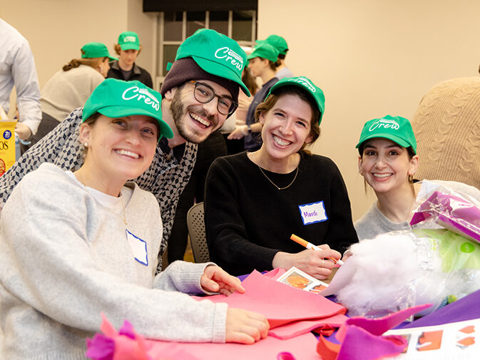 Group of young adults in green hats at a volunteer event doing a craft, smiling.