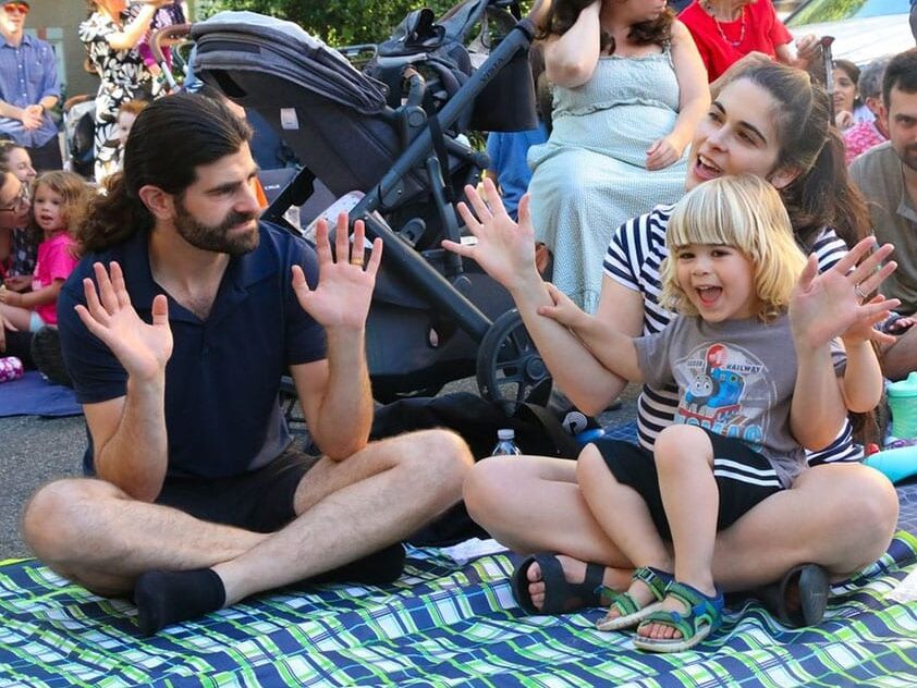 A family and their toddler sit on a picnic blanket at an outdoor event