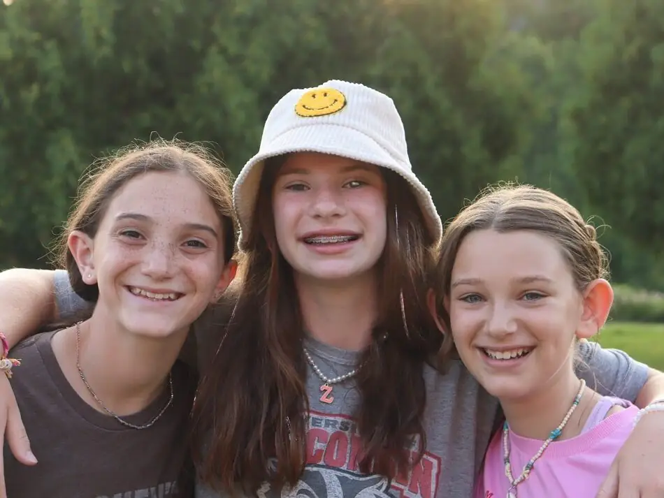 Close up of three teen girls smiling faces with arms around each other shoulders outside with trees in background.