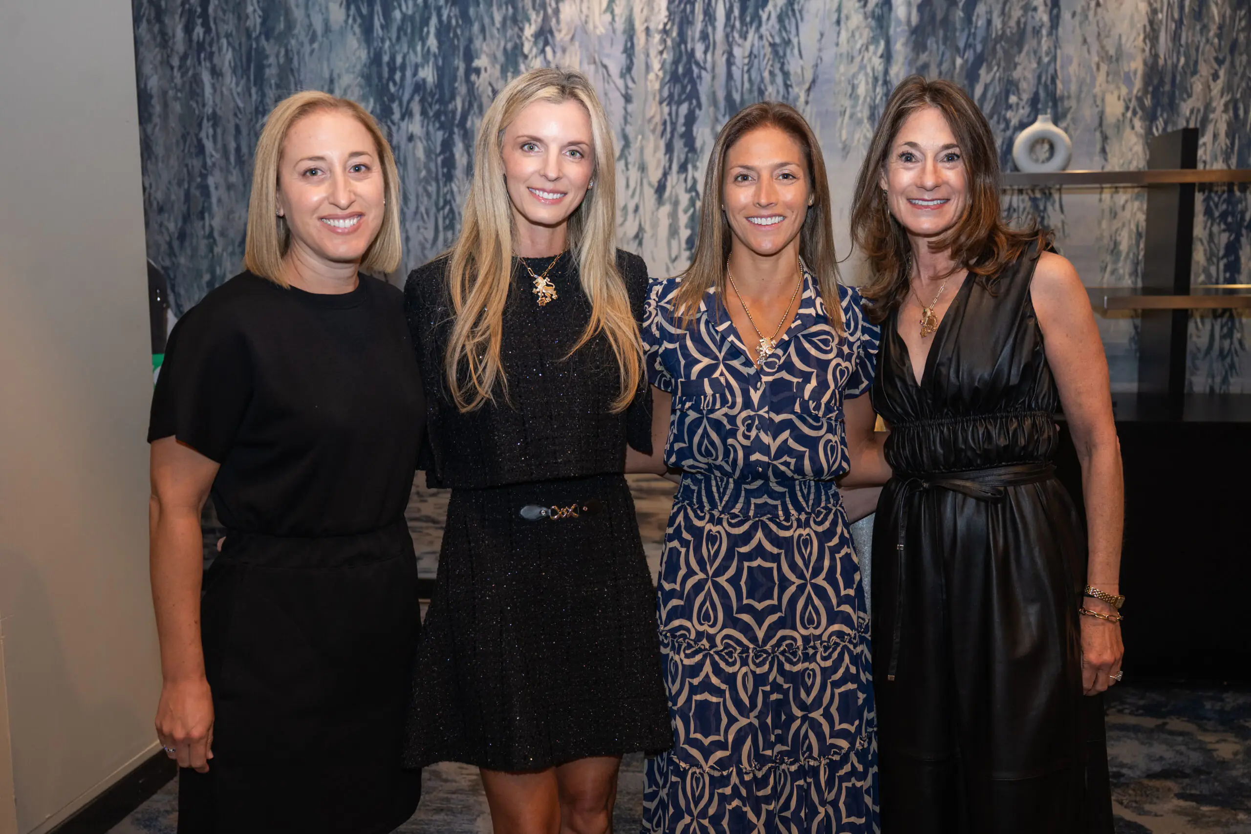 Four women standing side by side in front of a blue and white abstract background, dressed in various styles of black and patterned dresses.