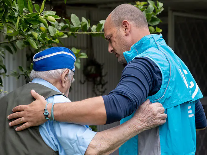 Two individuals outdoors, one wearing a blue cap and light blue shirt being assisted by a JDC case worker in a blue vest with white markings, with greenery in the background.