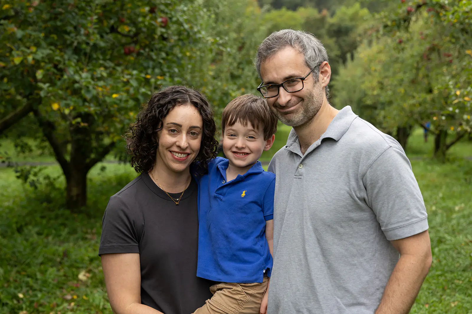 Mother, father, and son all smile in an apple orchard.